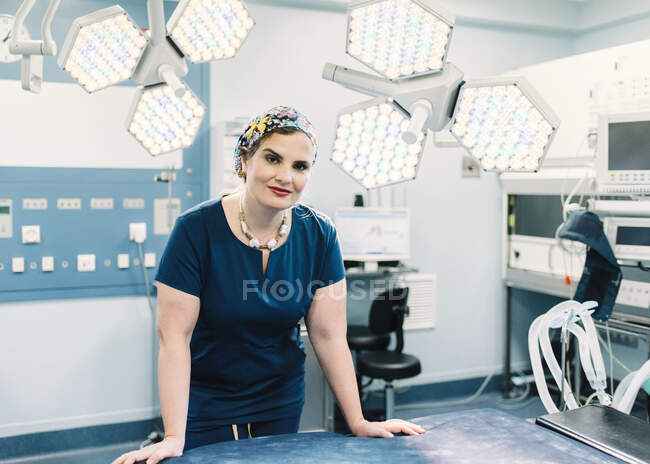 Доросла жінка в медичній формі дивиться на камеру, стоячи біля ламп в сучасному операційному театрі — стокове фото
