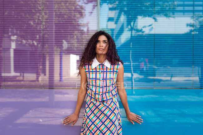 Sensual jovem etnia feminina em vestido colorido inclinado na parede de vidro de cor azul e rosa olhando para cima — Fotografia de Stock