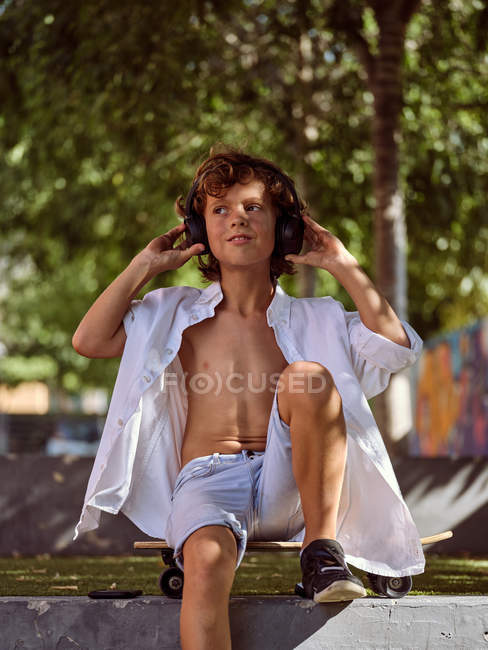 Niño casual pensativo en auriculares sentados en el monopatín mientras se relaja en el parque de skate en el soleado día de verano y mirando hacia otro lado - foto de stock
