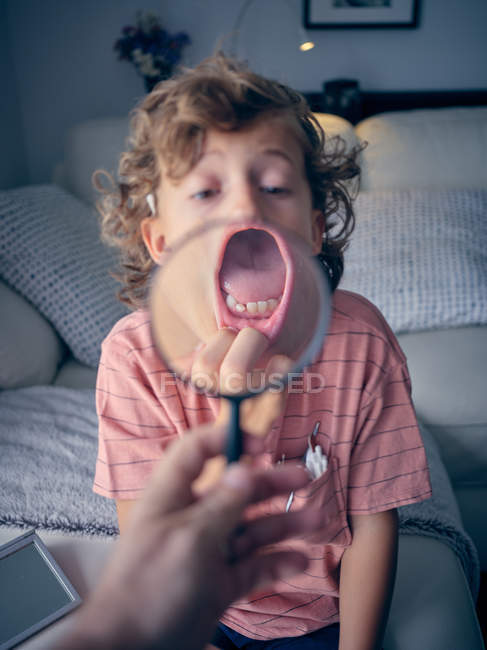 Surprised curly child showing tooth with mouth wide open pulling lower lip while person holding magnifying glass in room — Stock Photo