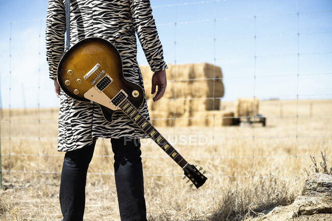 Crop vista posteriore di musicista elegante in zebra cappotto stampato con chitarra in piedi davanti recinzione filo spinato e guardando pagliai secchi in campo — Foto stock