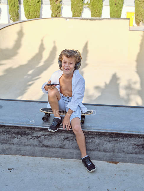 Niño con auriculares y el uso de teléfonos inteligentes mientras está sentado en el monopatín en el parque de skate urbano soleado - foto de stock