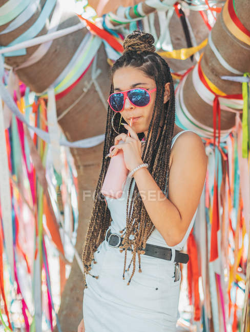 Стильна молода жінка в сонцезахисних окулярах п'є коктейль під час зовнішнього фестивалю і дивиться на камеру — стокове фото
