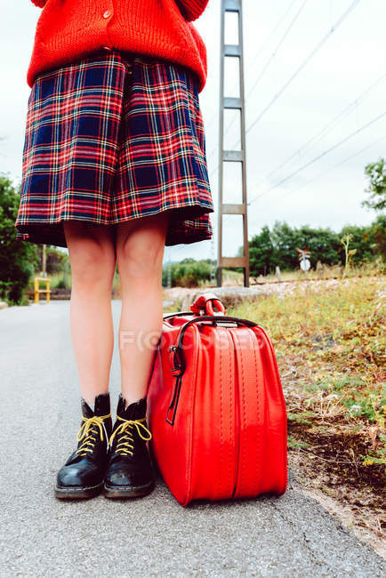 Жінка в стильних чоботях з червоною валізою, що стоїть на дорозі — стокове фото