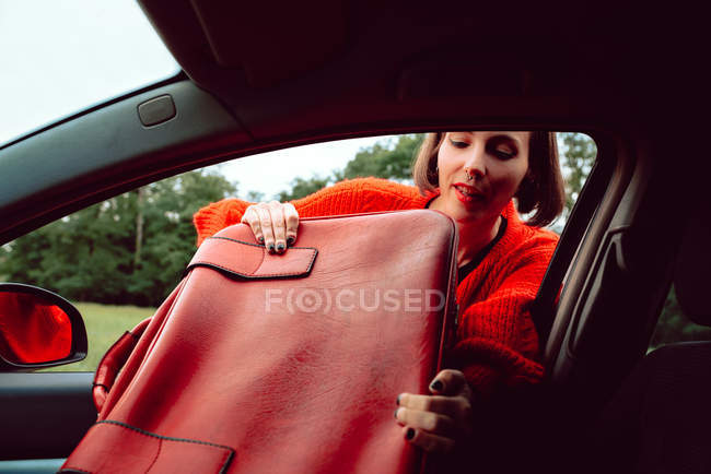 Donna mettendo valigia rossa vintage nel sedile anteriore della macchina attraverso il finestrino — Foto stock