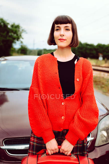 Affascinante donna attraente con capelli eleganti in maglione rosso con valigia rossa in attesa di viaggio in auto dietro sulla strada — Foto stock
