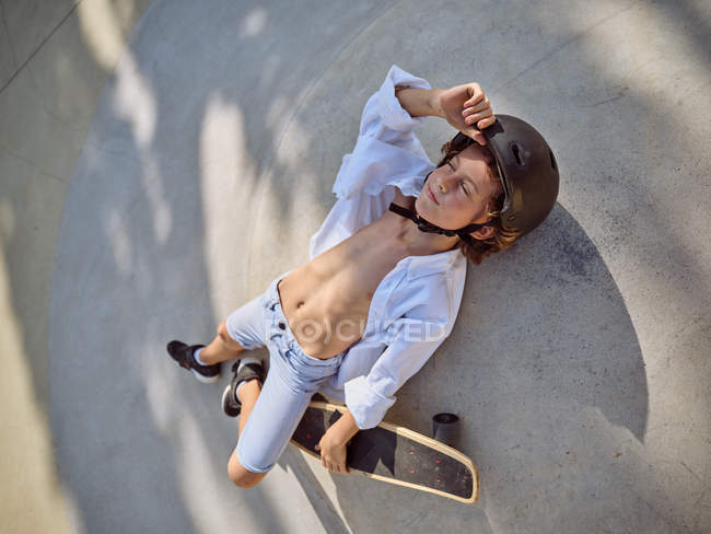 View from above of child in helmet lying down with eyes closed and chilling on ground in skatepark with shadows — Stock Photo