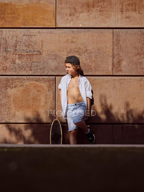 Хлопчик у білій сорочці стоїть, спираючись на стіну зі скейтбордом на сонячному світлі, дивлячись далеко — стокове фото
