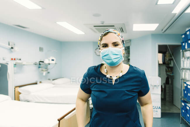 Female medic in blue uniform and protective mask standing in ward with empty beds and looking at camera — Stock Photo