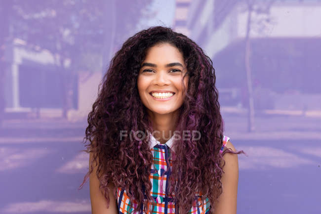 Retrato de la encantadora joven étnica con el pelo rizado mirando a la cámara contra la pared de vidrio púrpura - foto de stock