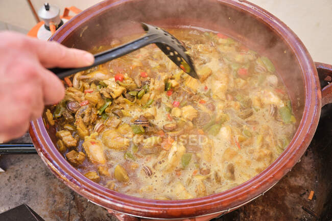 Crop anonymous standing human putting skimmer in large clay pot with cooking dish of chicken rice assorted vegetables and snails — Stock Photo