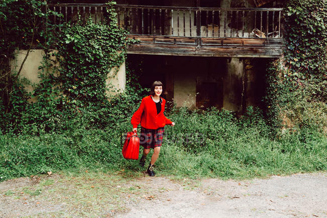 Молодая женщина в красном с большим красным чемоданом бегает в сельской местности против заброшенного дома — стоковое фото