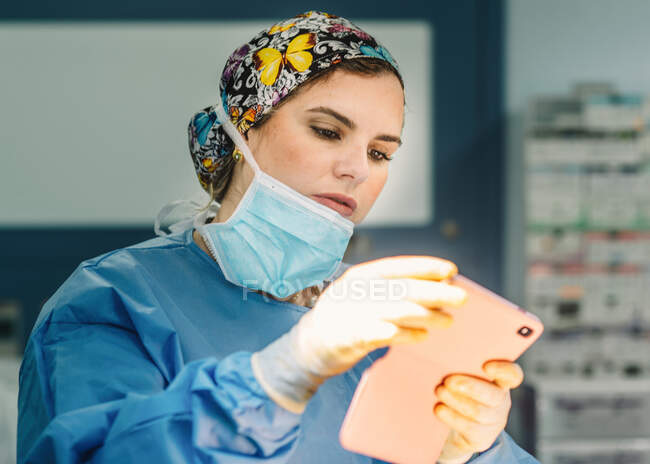 Красива жінка в хірургічній сукні та масці фотографує та використовує мобільний телефон під час роботи — стокове фото