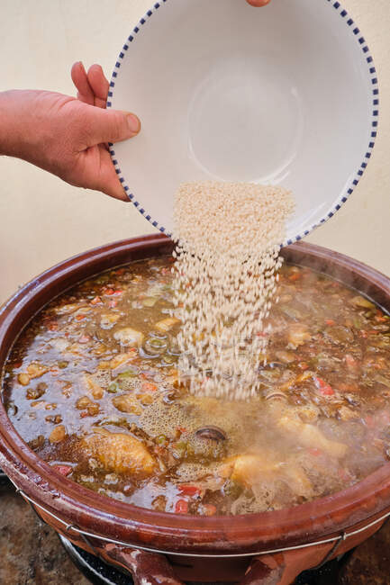 Cortar las manos de un humano anónimo vertiendo arroz en una olla grande de barro con un plato caliente de verduras de pollo y caracoles de Borgoña - foto de stock
