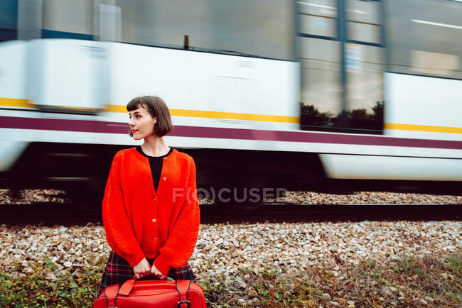 Стильная женщина в красном свитере с красным чемоданом, смотрящая вперед, в то время как скоростной поезд едет по железной дороге за спиной — стоковое фото