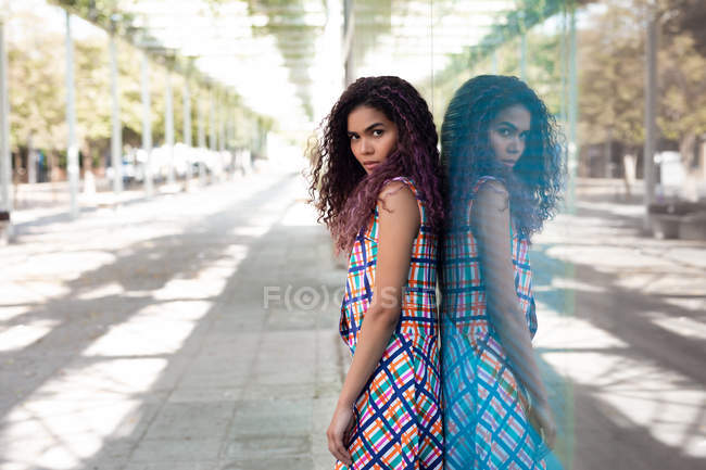 Seitenansicht einer jungen ethnischen Frau, die sich gegen eine Glaswand lehnt und auf der Straße reflektiert in die Kamera blickt — Stockfoto