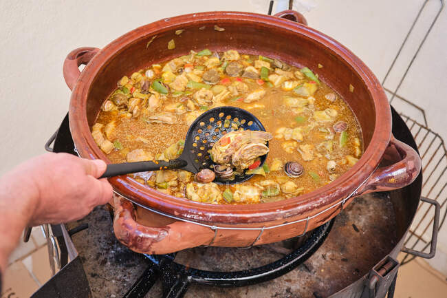 Crop anonyme debout humain mettre écumoire dans une grande casserole en argile avec plat de cuisson de riz de poulet assortiment de légumes et d'escargots — Photo de stock