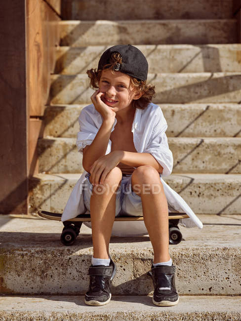 Cheerful boy sitting on skateboard on stairs in skatepark on sunny summer daytime — Stock Photo