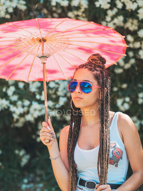 Slim young woman in summer outfit and sunglasses with umbrella standing near blooming trees — Stock Photo