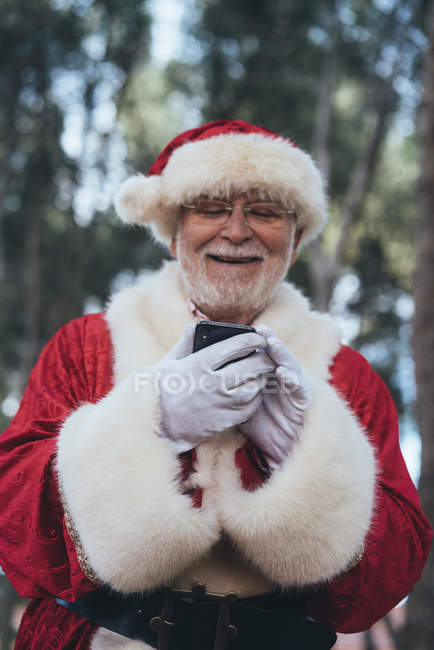 Uomo gioioso in costume di Babbo Natale utilizzando il moderno telefono cellulare su sfondo natura offuscata — Foto stock