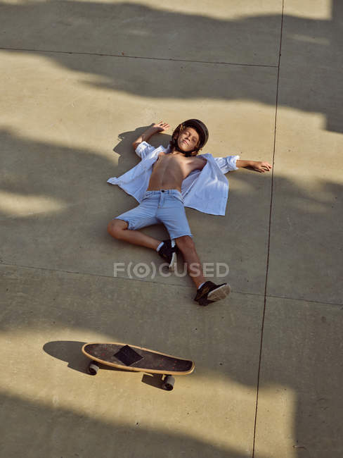 Blick von oben auf ein Kind im Helm, das mit geschlossenen Augen auf dem Boden im Skatepark mit Schatten liegt — Stockfoto