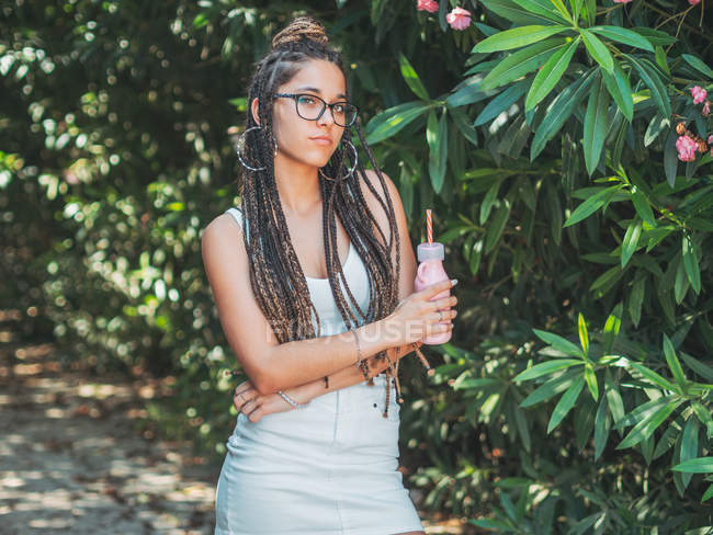 Trendige junge Frau in Brille mit Dreadlocks trinkt Cocktail und blickt im Garten in die Kamera — Stockfoto