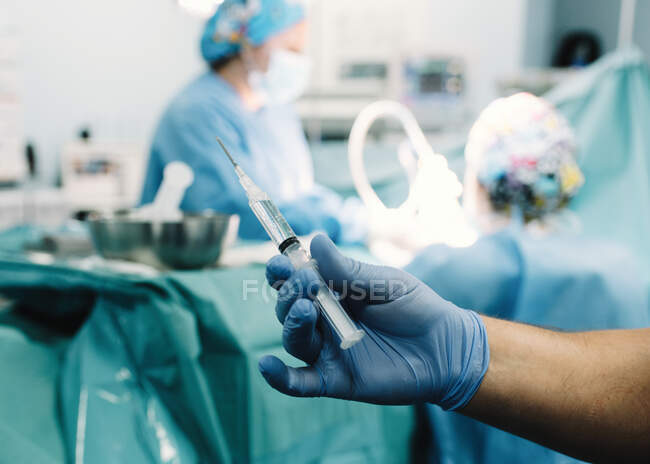 Mano de guante de cultivo con inyección preparada y cirujanos desenfocados en el trabajo en quirófano - foto de stock