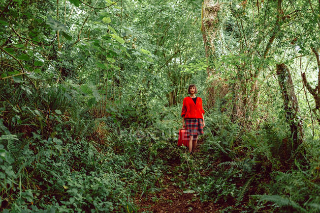 Donna in rosso con grande valigia rossa passeggiando nella foresta verde — Foto stock
