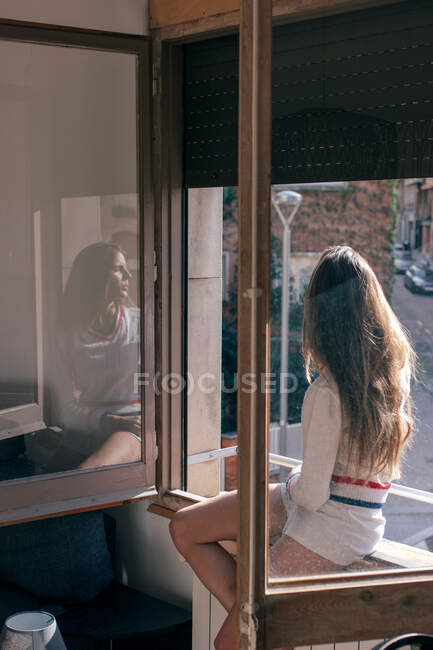 Vista laterale di donna snella seduta sul davanzale della finestra in appartamento e guardando lontano in luce morbida — Foto stock