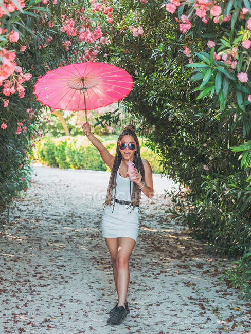 Весела струнка молода жінка в літньому вбранні і сонцезахисних окулярах з парасолькою, що п'є напій біля квітучих дерев — стокове фото
