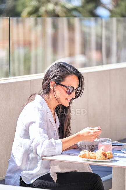 Довольно дружелюбная дама в солнечных очках сидит за столом на открытой солнечной террасе и ест розовый йогурт в ожидании партнера — стоковое фото