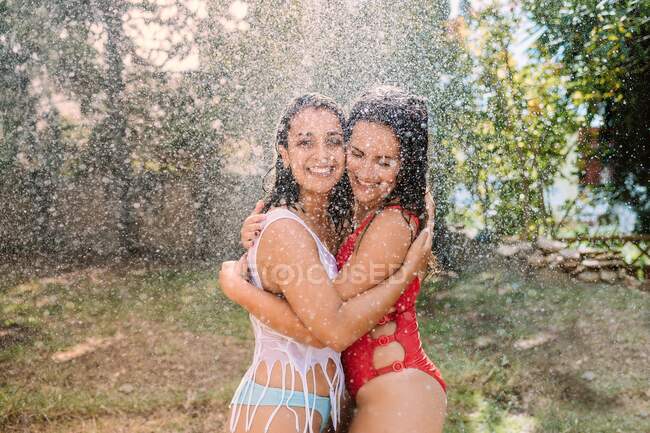 Cheerful beautiful girlfriends in swimsuits embracing happily while standing in splashing fountain of water drops in garden — Stock Photo