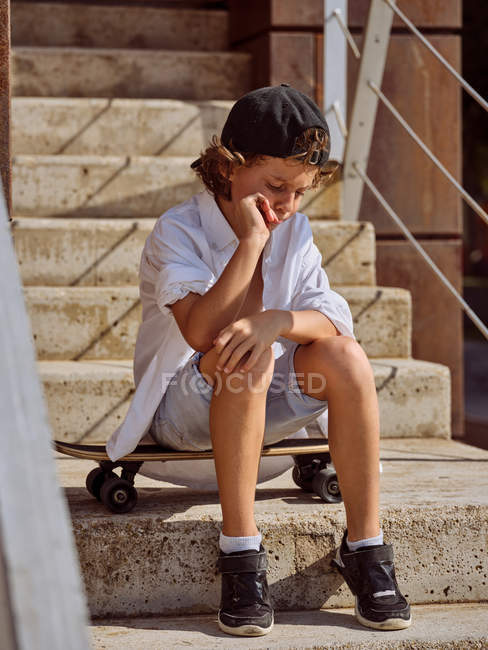 Веселий хлопчик сидить на скейті на сходах у скейтпарку в сонячне літо, дивлячись вниз — стокове фото