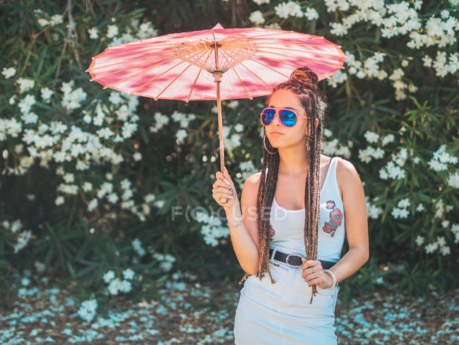 Schlanke junge Frau im Sommeroutfit und Sonnenbrille mit Regenschirm, die neben blühenden Bäumen steht — Stockfoto