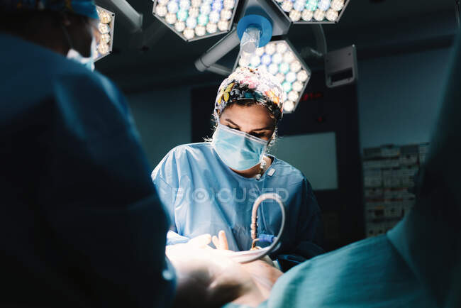 Da sotto grave giovane medico in maschera protettiva e cap facendo chirurgia con strumenti e crop nurse — Foto stock