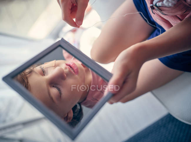 Відбиття в квадратному дзеркалі обличчя дитини з бинтом на щоці вивчає молочний зуб з відкритим ротом в кімнаті — стокове фото