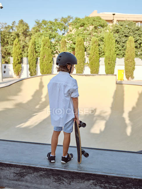 Вид сзади на мальчика в защитном шлеме и катание на скейтборде на рампе в скейтпарке — стоковое фото