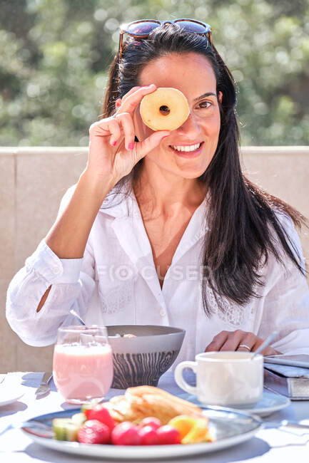 Frau sitzt am Tisch mit serviertem Frühstück auf offener sonnenbeschienener Terrasse, während sie Donut hält und vorführt und lacht, während sie auf verschwommenem Hintergrund in die Kamera blickt — Stockfoto
