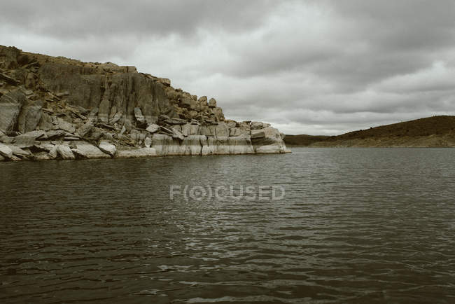 Gloomy seascape of rocky cliff and water with shallow waves reflecting gray sky — Stock Photo