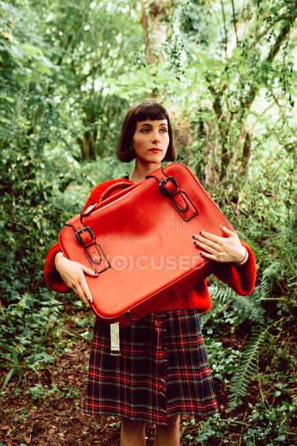 Женщина в красном с большим красным чемоданом гуляет по зеленому лесу — стоковое фото