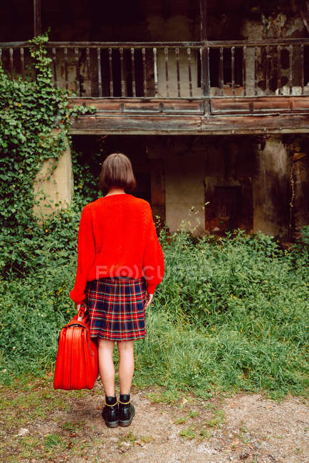 Женщина в красном с большим винтажным красным чемоданом стоит перед заброшенным домом в сельской местности — стоковое фото