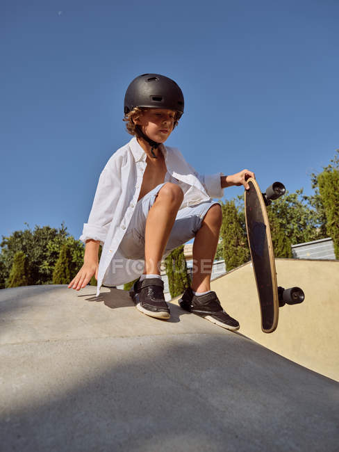 Dal basso del ragazzo felice con il casco che tiene lo skateboard seduto sulla rampa contro il cielo blu — Foto stock