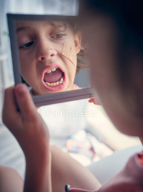 Reflexion im quadratischen Spiegel des Gesichts des Kindes mit Binde auf der Wange beim Studium des Milchzahns mit offenem Mund im Zimmer — Stockfoto