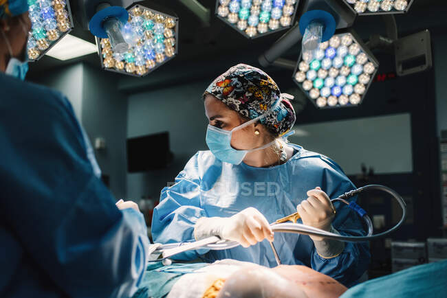 Jovem médico grave com máscara protetora e tampa fazendo cirurgia com instrumentos e enfermeira de colheita — Fotografia de Stock