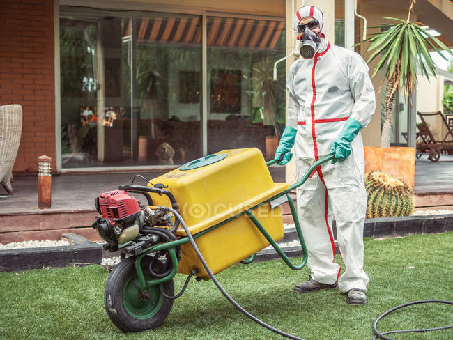 Fumigator in white uniform, respiratory mask and green gloves holding yellow tank on wheel in yard and looking at camera — Stock Photo