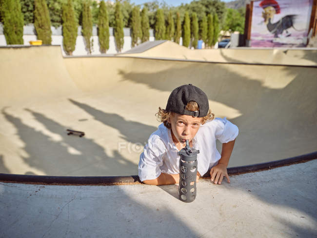 Cooler kleiner Junge mit Schirmmütze hängt an Skaterrampe und trinkt Wasser aus Sportflasche mit Strohhalm, der in die Kamera schaut — Stockfoto