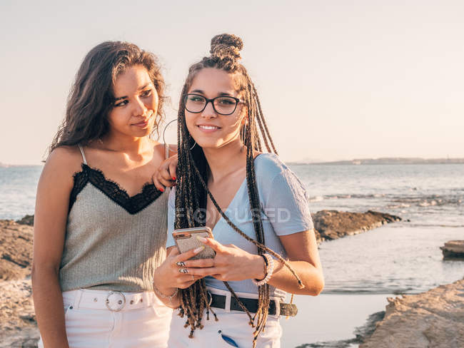 Улыбающиеся модные молодые женщины с помощью смартфона на пляже — стоковое фото