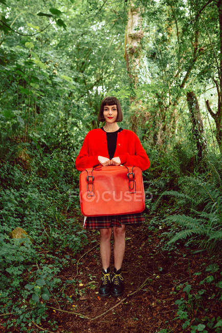 Mulher de vermelho com mala vermelha vintage em pé na floresta — Fotografia de Stock