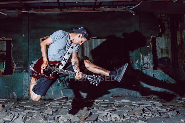 Музыкант-мужчина экспрессивно прыгает, играя на электрогитаре в темном полуразрушенном зале — стоковое фото