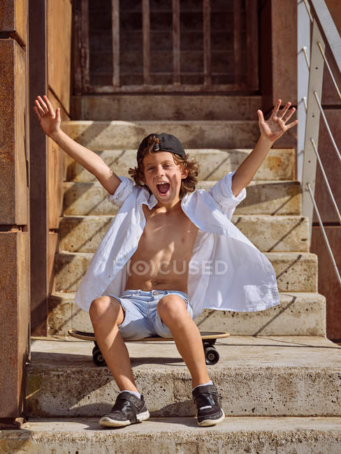 Веселий хлопчик сидить на скейтборді на сходах у скейтпарку на сонячне літо дивлячись в камеру — стокове фото